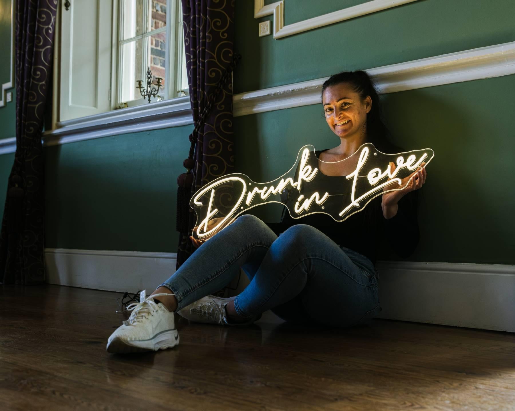 Person holding a neon sign with 'Drunk in Love' in a room with green walls and a window.
